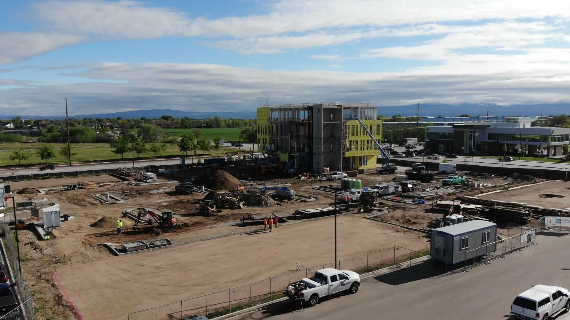 Aerial view of a building going vertical at Discovery Meridian, with workers in orange vests on a survey-marked pad in the foreground and an excavator working — April 22, 2026