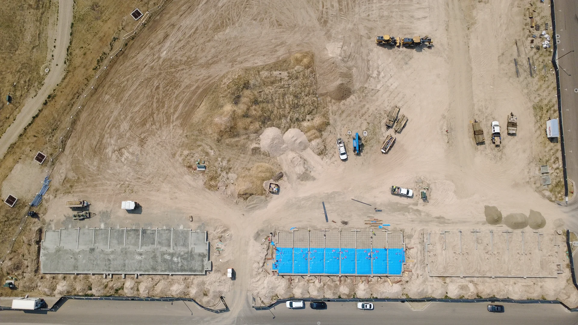 Aerial view of the Arête flats foundations in August 2026 — rows of formwork and rebar under blue tarps, with a poured concrete slab section and construction equipment on site